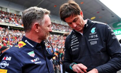Two men in dark blue racing team jackets talk in a crowded stadium, one in profile facing left and the other looking down, with fans in the background.