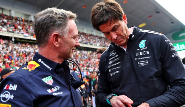 Two men in dark blue racing team jackets talk in a crowded stadium, one in profile facing left and the other looking down, with fans in the background.