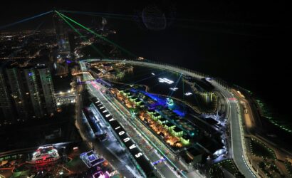 Aerial night view of a brightly lit city waterfront with tall buildings, curved roadway, and green laser beams crisscrossing the sky beside a dotted illuminated sphere.