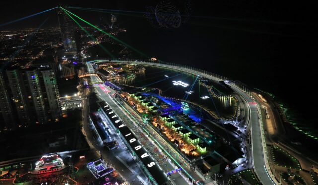 Aerial night view of a brightly lit city waterfront with tall buildings, curved roadway, and green laser beams crisscrossing the sky beside a dotted illuminated sphere.