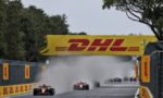 Formula 1 cars race down a straight under a large yellow DHL banner, kicking up dust and smoke behind them.