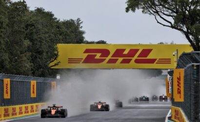 Formula 1 cars race down a straight under a large yellow DHL banner, kicking up dust and smoke behind them.
