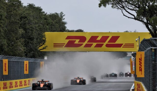 Formula 1 cars race down a straight under a large yellow DHL banner, kicking up dust and smoke behind them.