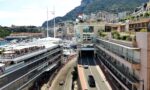 Aerial view of Monaco harbour with many yachts, bordered by modern buildings and a green hillside cityscape in the background.