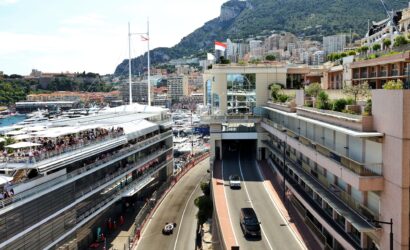 Aerial view of Monaco harbour with many yachts, bordered by modern buildings and a green hillside cityscape in the background.