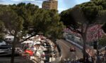 Crowded Monaco street circuit with a red race car on track, grandstands, boats, and trees framing the scene.