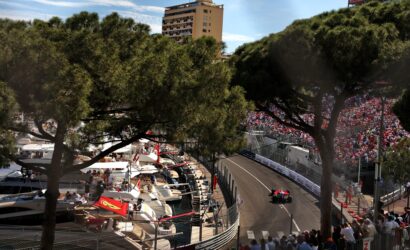 Crowded Monaco street circuit with a red race car on track, grandstands, boats, and trees framing the scene.