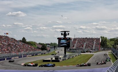 Crowded Formula 1 race on a banked track with multiple cars rounding a long curve, grandstands full of spectators, and sponsor banners along the walls.