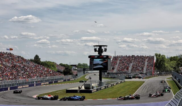 Crowded Formula 1 race on a banked track with multiple cars rounding a long curve, grandstands full of spectators, and sponsor banners along the walls.