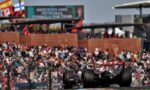 Formula 1 car racing past a crowded grandstand with flags and banners in the background (Mercedes AMG Petronas signage)