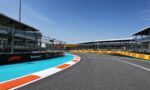 Curved Formula 1 race track with blue runoff, orange-white curbs, safety barriers, and grandstands along the pit buildings under a clear blue sky.