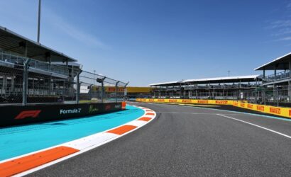 Curved Formula 1 race track with blue runoff, orange-white curbs, safety barriers, and grandstands along the pit buildings under a clear blue sky.