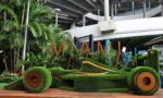 Outdoor urban garden installation with manicured green turf sculptures and large orange letters spelling MIAMI against a concrete overpass.