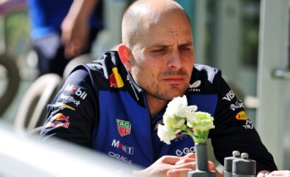 Male race car driver in a blue sponsor-laden suit, looking down while holding white flowers.