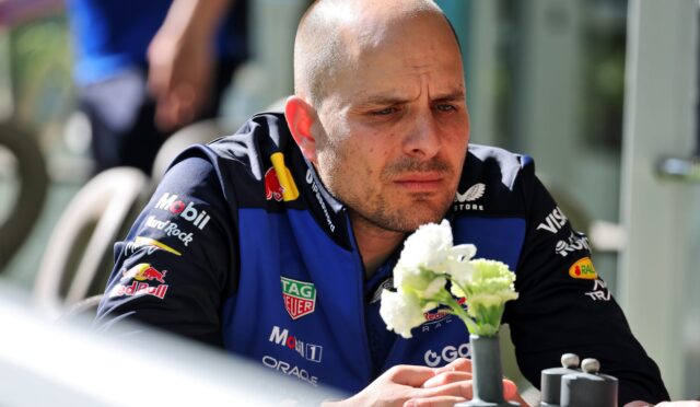 Male race car driver in a blue sponsor-laden suit, looking down while holding white flowers.