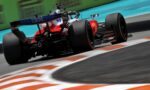 Rear three-quarter view of a red Formula 1 race car speeding along a track, with motion blur and orange-and-white curbs in the foreground.