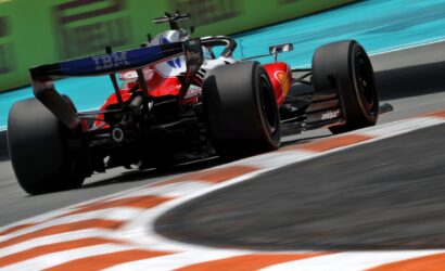 Rear three-quarter view of a red Formula 1 race car speeding along a track, with motion blur and orange-and-white curbs in the foreground.