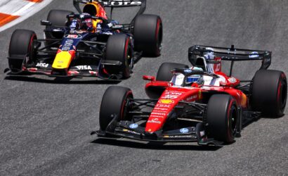 Three Formula 1 cars race in a tight pack on a track, led by a red Ferrari in the foreground.