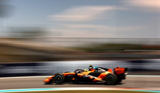 Orange Formula 1 car racing at high speed on a track, bumper-to-bumper motion blur in the background showing speed.