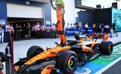 Pit lane scene with a Formula 1 driver in an orange suit standing on his race car, raising a hand in victory beside a brightly colored Renault-style car and team members in the background.