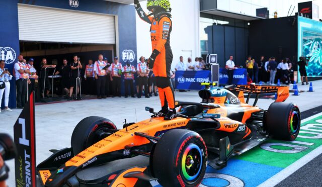 Pit lane scene with a Formula 1 driver in an orange suit standing on his race car, raising a hand in victory beside a brightly colored Renault-style car and team members in the background.
