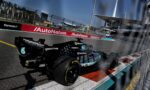 Black Formula 1 car speeding on a race track with sponsor logos (Petronas, Microsoft) visible and blurred fencing in the foreground; grandstands and pit buildings in distance.