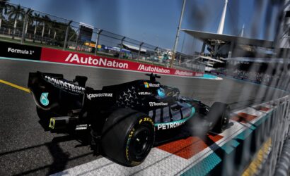 Black Formula 1 car speeding on a race track with sponsor logos (Petronas, Microsoft) visible and blurred fencing in the foreground; grandstands and pit buildings in distance.