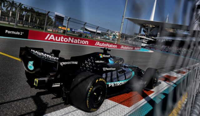 Black Formula 1 car speeding on a race track with sponsor logos (Petronas, Microsoft) visible and blurred fencing in the foreground; grandstands and pit buildings in distance.