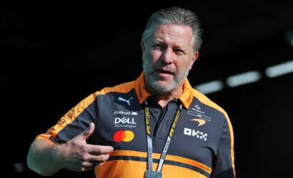 Man in an orange and black polo with sponsor logos, wearing a lanyard, gesturing as he speaks at an event.