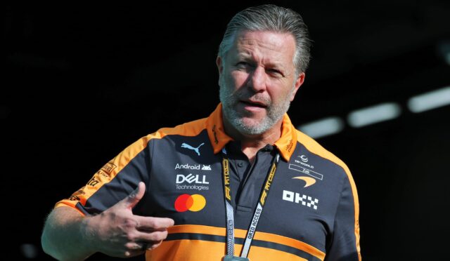 Man in an orange and black polo with sponsor logos, wearing a lanyard, gesturing as he speaks at an event.