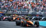 Formula 1 cars sprint off the grid at the start of a race, with a packed crowd cheering in the stands behind a barrier.