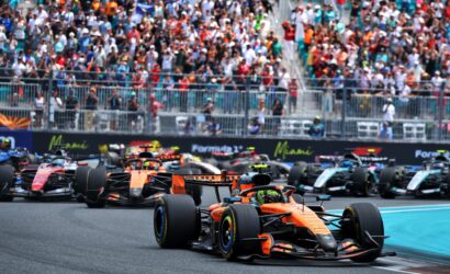 Formula 1 cars sprint off the grid at the start of a race, with a packed crowd cheering in the stands behind a barrier.