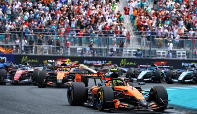 Formula 1 cars sprint off the grid at the start of a race, with a packed crowd cheering in the stands behind a barrier.