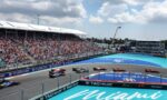 Crowded grandstands overlook a Formula 1 race on the Miami street circuit, with several cars racing around a curve and sponsor banners along the barriers.