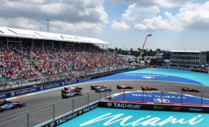 Crowded grandstands overlook a Formula 1 race on the Miami street circuit, with several cars racing around a curve and sponsor banners along the barriers.