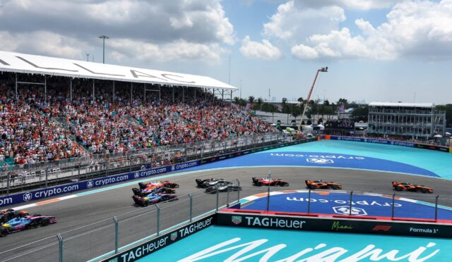 Crowded grandstands overlook a Formula 1 race on the Miami street circuit, with several cars racing around a curve and sponsor banners along the barriers.