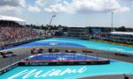 Formula 1 race on a turquoise Miami track with colorful cars and a crowded grandstand on the left.