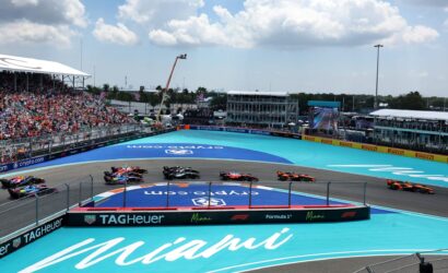 Formula 1 race on a turquoise Miami track with colorful cars and a crowded grandstand on the left.