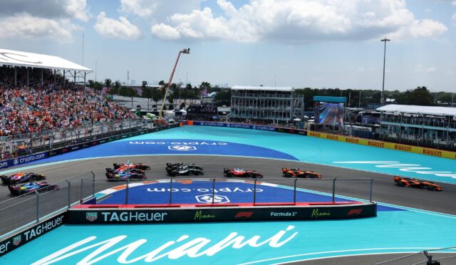 Formula 1 race on a turquoise Miami track with colorful cars and a crowded grandstand on the left.