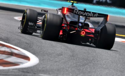 Rear view of a Formula 1-style race car taking a corner, red taillights illuminated, Revolut branding on the rear wing under bright track lighting.