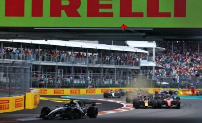 Formula 1 cars race on a street circuit with a crowd watching from elevated grandstands and bright Pirelli barriers nearby.