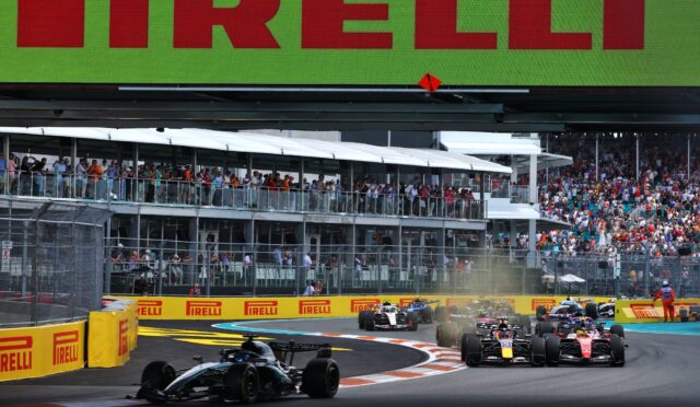 Formula 1 cars race on a street circuit with a crowd watching from elevated grandstands and bright Pirelli barriers nearby.