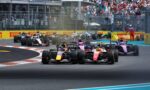 Formula 1 cars race side by side into a turn, with a crowded grandstand and safety barriers in the background.