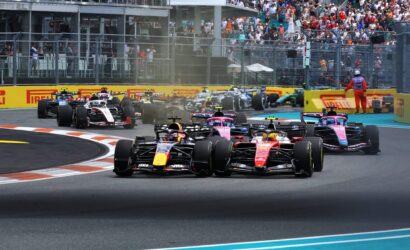 Formula 1 cars race side by side into a turn, with a crowded grandstand and safety barriers in the background.