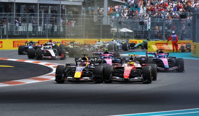 Formula 1 cars race side by side into a turn, with a crowded grandstand and safety barriers in the background.