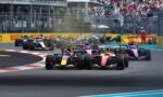 Row of Formula 1 cars on track at the start of a race, smoke rising as they accelerate, crowded grandstands in the background.