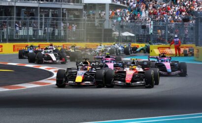 Row of Formula 1 cars on track at the start of a race, smoke rising as they accelerate, crowded grandstands in the background.