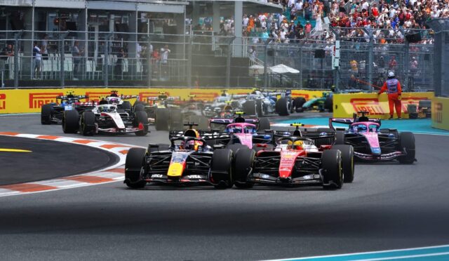 Row of Formula 1 cars on track at the start of a race, smoke rising as they accelerate, crowded grandstands in the background.