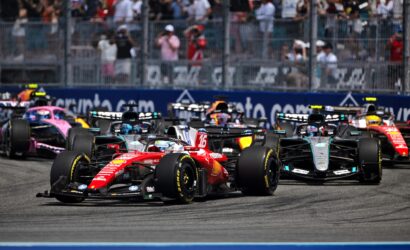 Race cars start a Grand Prix on a track with a cheering grandstand in the background, front red car leading the pack.