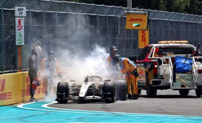 Formula 1 car halted after crash with smoke; safety crew and a fire extinguisher on-track near yellow barriers and a DHL sign above the fence.